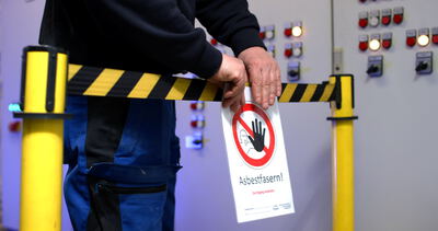 Arbeiter bringt Warnschild vor Asbestfasern an einer Absperrung in einem industriellen Schaltschrankraum an