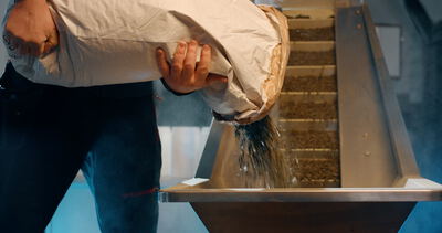 Arbeiter schüttet dunkles Schüttgut aus einem großen Papiersack in den Einfülltrichter einer industriellen Förderanlage