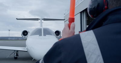 Einweiser mit Leuchtstab dirigiert einen weißen Privatjet auf dem Rollfeld vor einer Flugzeughalle