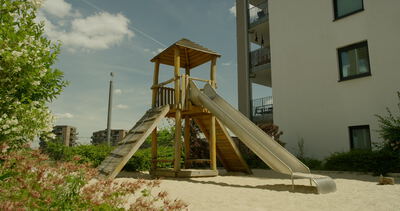 Holzturm mit Rutsche und Klettermöglichkeit auf einem Sandspielplatz vor modernen Wohngebäuden an einem sonnigen Tag
