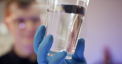 Laborant prüft Wasserprobe in einem Glas mit Schutzbrille und Handschuhen: Labor, Wasserprobe