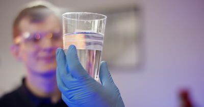 Laborant prüft Wasserprobe in einem Glas mit Schutzbrille und Handschuhen
