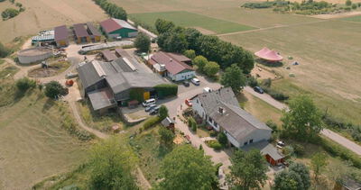 Luftaufnahme eines weitläufigen Bauernhofs mit Stallungen, Wohnhäusern und angrenzenden Feldern