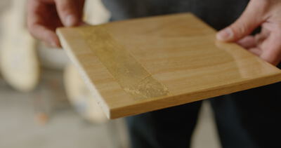 Nahaufnahme von Händen die eine lackierte Holzplatte mit einem aufgebrachten Streifen Blattgold zur Prüfung halten