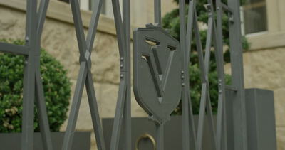Nahaufnahme von einem grauen Metalltor mit einem wappenförmigen V-Logo vor einer Sandsteinfassade Nahaufnahme von einem grauen Metalltor mit einem wappenförmigen V-Logo vor einer Sandsteinfassade, Gitter, Metallgitter und