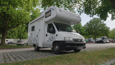 Wohnmobil steht in Dreiviertelansicht schräg von vorn auf einem schattigen Rastplatz: Wohnmobil Dreiviertelansicht, Reisemobil auf Stellplatz