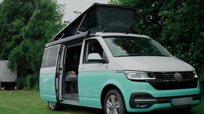 Türkiser Campingbus mit Aufstelldach steht auf einer Wiese am Waldrand