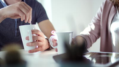 Person rührt Kaffee in einer weißen Tasse am hellen Tisch