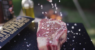 Grobe Salzflocken fallen auf ein rohes Steak mit Pfeffer vor dem Grillen