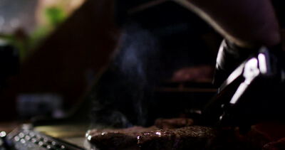 Behandschuhte Hand grillt Fleisch mit Zange über Flammen auf dem Grillrost