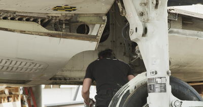 Person arbeitet unter einem Flugzeugrumpf neben dem Fahrwerk