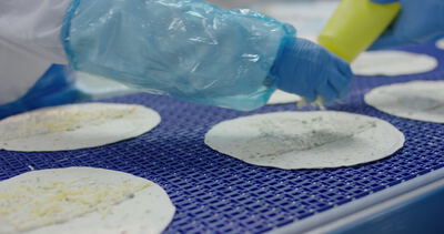 Käse wird am Förderband auf runde Teigfladen aufgelegt