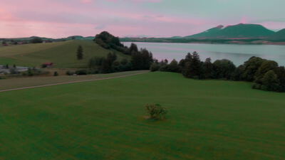 Drohnenflug über grüne Voralpenwiesen mit Bergsee im rosa Morgenlicht
