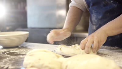 Bäcker in blauer Schürze drückt Teig auf heller Arbeitsplatte, Bäcker drückt Teig, blaue Schürze in der Bäckerei und