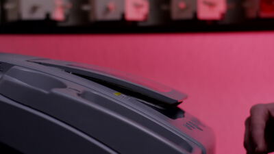 Person öffnet den Deckel einer grauen Maschine vor rosa Wand