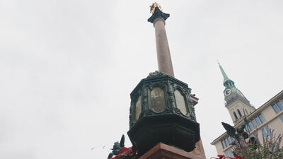 Mariensäule mit goldener Figur ragt vor einem Kirchturm empor