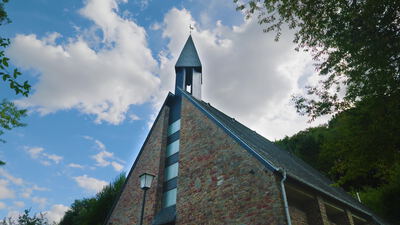 Grüne Baumkronen schieben sich vor den Giebel einer Steinkirche, Kirche zwischen Bäumen, Kirchturmspitze und Wetterhahn als