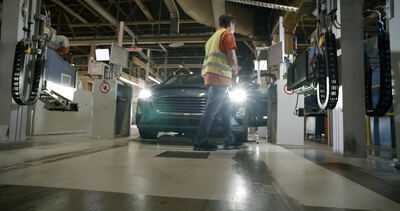 Werker in Warnweste geht an einem Fahrzeug in der Fertigungshalle vorbei