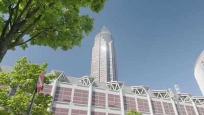 Der Messeturm in Frankfurt ragt hinter grünen Bäumen in den blauen Himmel