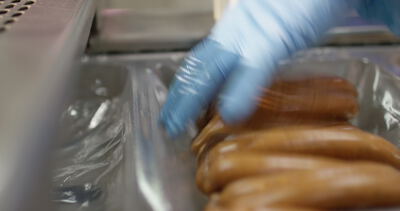 Hand mit blauem Handschuh legt Würstchen in eine Verpackungsschale