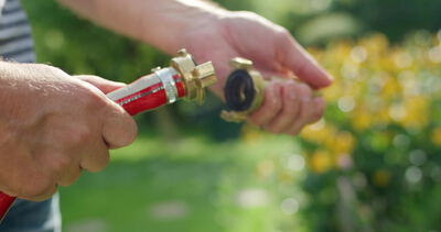 Hände verbinden eine Messingkupplung an einem roten Gartenschlauch im Garten
