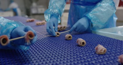 Arbeiter stecken gefüllte Baconröllchen auf Spieße an einem blauen Förderband