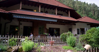Gasthaus mit Terrasse und rotem Ziegeldach am Rand eines Waldes