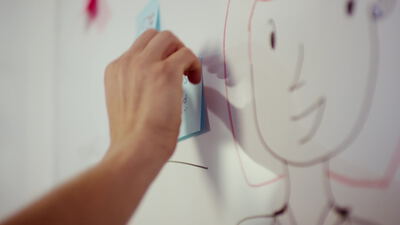 Hand klebt einen blauen Notizzettel an ein Whiteboard