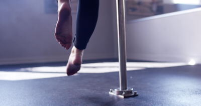 Barfüßige Person turnt an vertikaler Stange im hellen Trainingsraum