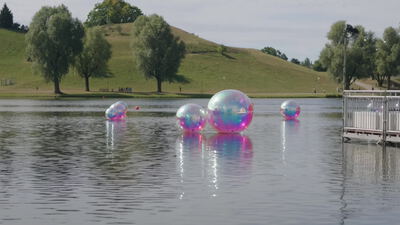 Bunte Kugeln schwimmen auf einem See vor einem grünen Hügel: Kugeln auf dem See, grüner Hügel im Park