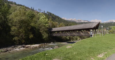 Überdachte Holzbrücke über einen Gebirgsbach zwischen Wiese und Wald